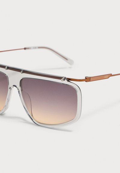 Lunettes de soleil rectangulaires avec montures transparentes, verres marron dégradés et fines branches métalliques marron avec un petit logo sur le côté.