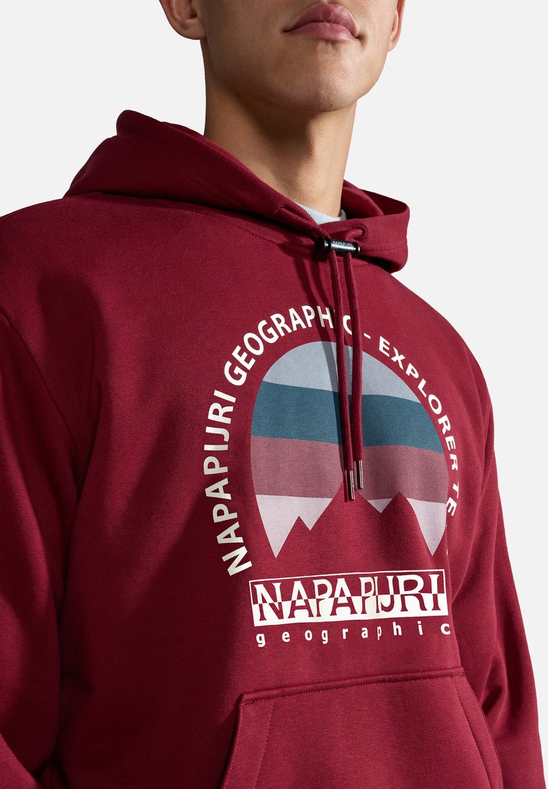 Mężczyzna w bordowej bluzie z kapturem z grafiką góry oraz napisem "Zespół Ekspedycji Napapijri" na piersi.