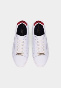 Par af hvide Tommy Hilfiger letvægts sneakers med hvide snørebånd, røde hælaccents og metalliske "TOMMY" snøretags, set oppefra.