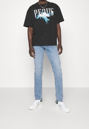 Man wearing black graphic t-shirt, light blue jeans, white sneakers, and a silver chain necklace, standing against a plain white background.