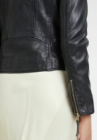 Giacca in pelle nera con design aderente, cuciture visibili e cerniera dorata al polso. Tessitura liscia con silhouette classica.