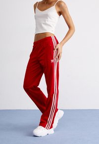 Person som bär vita sneakers, röda Adidas träningbyxor med vita ränder och en vit kort topp, poserar på en ljusblå golv.