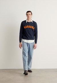 Suéter azul-marinho com a letra em laranja "BARBOUR", usado sobre uma camisa branca com colarinho, combinado com jeans azul claro e sapatos pretos.