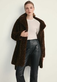 Brązowy płaszcz z futra syntetycznego w cętki lamparta z szerokim kołnierzem, noszony na jasnej bluzce i zestawiony z czarnymi skórzanymi spodniami. Gładka tekstura.