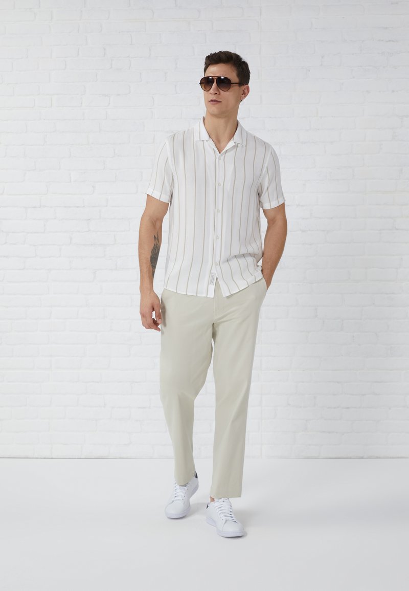 Chemise blanche à manches courtes avec des rayures verticales beiges, pantalon beige clair et baskets blanches ; mannequin portant des lunettes de soleil noires.