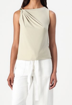 Haut beige sans manches avec tissu froncé et détail anneau doré sur l'épaule gauche, porté avec un pantalon blanc taille haute possédant des poches devant et une ceinture à nouer.