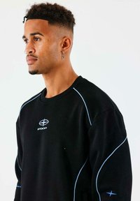Sudadera negra con acentos y costuras azules, que presenta un logo en el pecho. Tejido suave, cuello redondo y diseño de corte relajado.