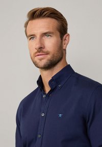 Marineblauwe, knoopsluiting shirt met een klein geborduurd motief op de borst. Voorzien van een gestructureerde kraag en een gladde, textuurstof.