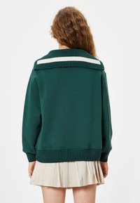 Sudadera verde oscura con un cuello acanalado de rayas blancas, usada sobre una falda plisada beige claro. Tela de algodón con un ajuste relajado.