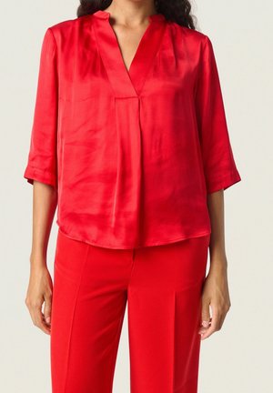 Kobieta ubrana w jasnoczerwoną, luźną satynową bluzkę z rękawami do łokcia oraz dopasowane czerwone spodnie.