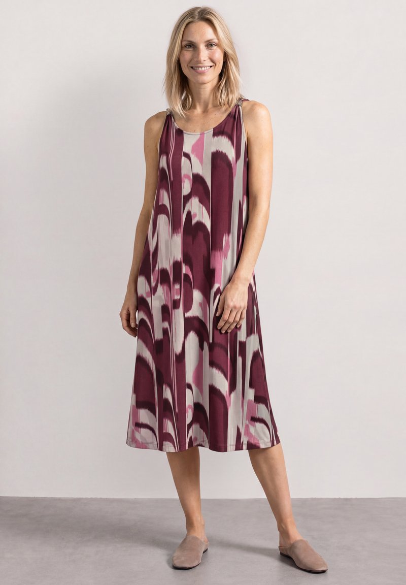 Femme aux cheveux blonds portant une robe sans manches longueur genoux avec un motif abstrait violet et rose et des chaussures beiges à enfiler, debout contre un mur blanc.