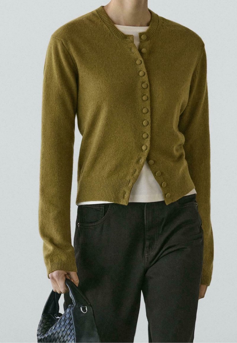 Personne portant un cardigan boutonné vert olive sur un haut blanc et un pantalon noir, tenant un sac à main noir tressé dans une main.