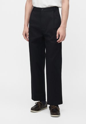 Persona che indossa pantaloni neri a gamba larga, top bianco, calze scure e scarpe da barca marrone scuro, in piedi davanti a uno sfondo bianco.