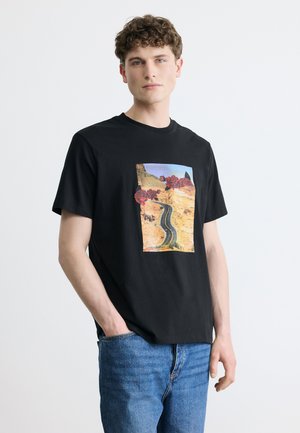 Młody mężczyzna o kręconych włosach, ubrany w czarny T-shirt z grafiką drogi na pustyni i czerwonych róż, w połączeniu z niebieskimi dżinsami, stojący na białym tle.