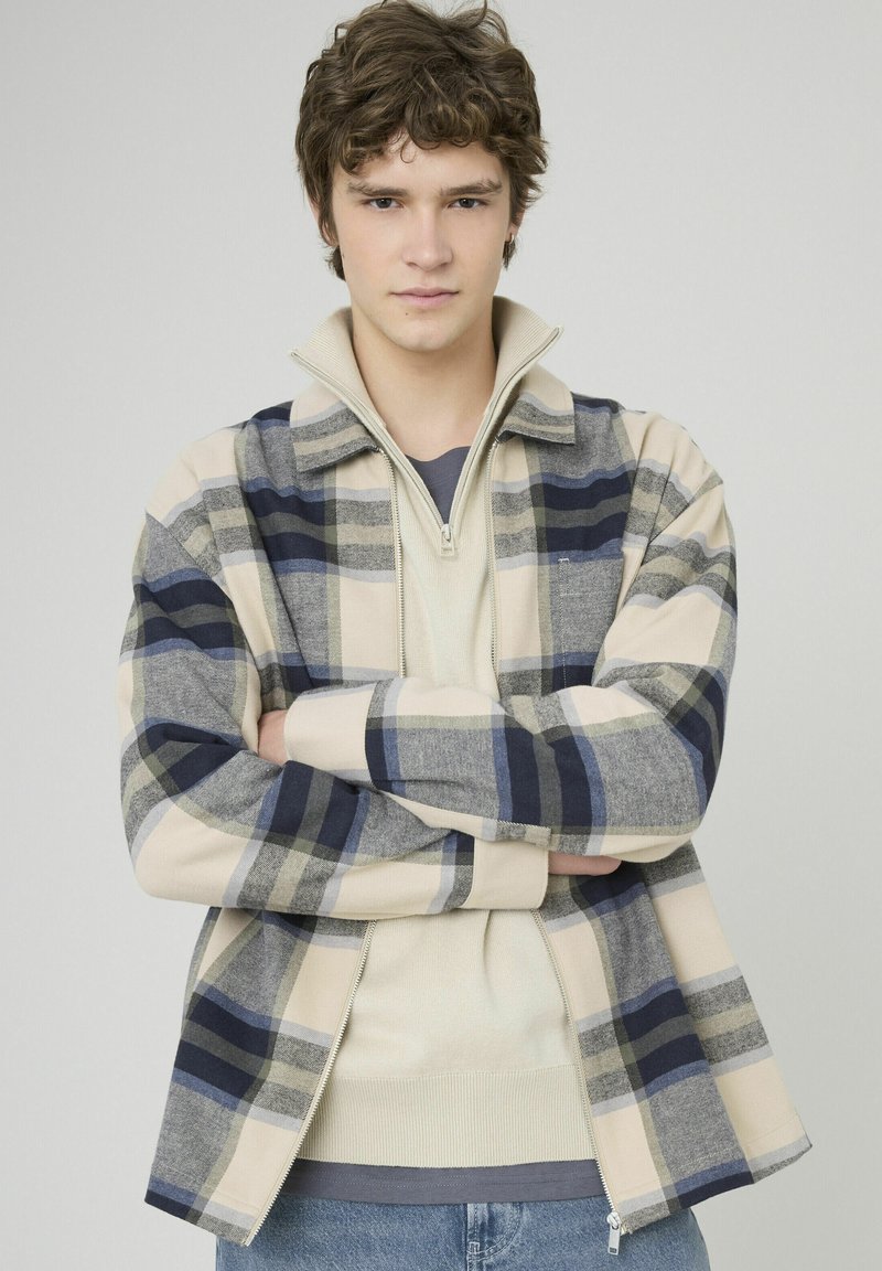 Jeune homme aux cheveux bouclés portant une veste à carreaux beige, marine et grise par-dessus un pull à col zippé crème, se tenant les bras croisés.