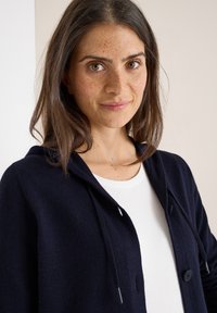Giovane donna con i capelli castani e lentiggini indossa un cardigan blu navy sopra una maglietta bianca, sorridendo dolcemente su uno sfondo chiaro.