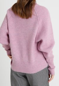 Suéter de punto acanalado en color rosa, con un corte holgado, que presenta mangas raglán y un cuello amplio, combinado con pantalones de patrón en blanco y negro.