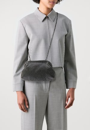 Mujer vestida con una camisa de manga larga a cuadros en blanco y negro y pantalones, llevando un pequeño bolso negro adornado con strass y con una correa de cadena.