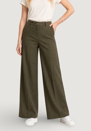 Femme portant un pantalon large taille haute vert olive, une chemise blanche à manches courtes, un bracelet argenté et des baskets blanches, debout avec une main dans la poche.