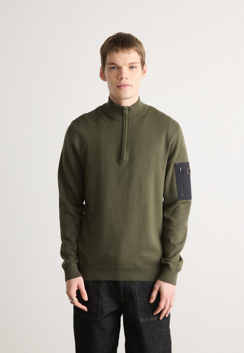 Olivengrüner Reißverschluss-Pullover mit hohem Kragen, langen Ärmeln und schwarzem aufgenähtem Taschen auf dem linken Arm. Weiches Strickmaterial und taillierte Passform.