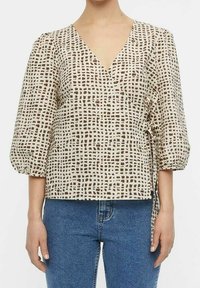 Top beige enroulé avec un décolleté en V, motif en grille texturé marron, manches bouffantes et un lien sur le côté, associé à un jean en denim bleu.