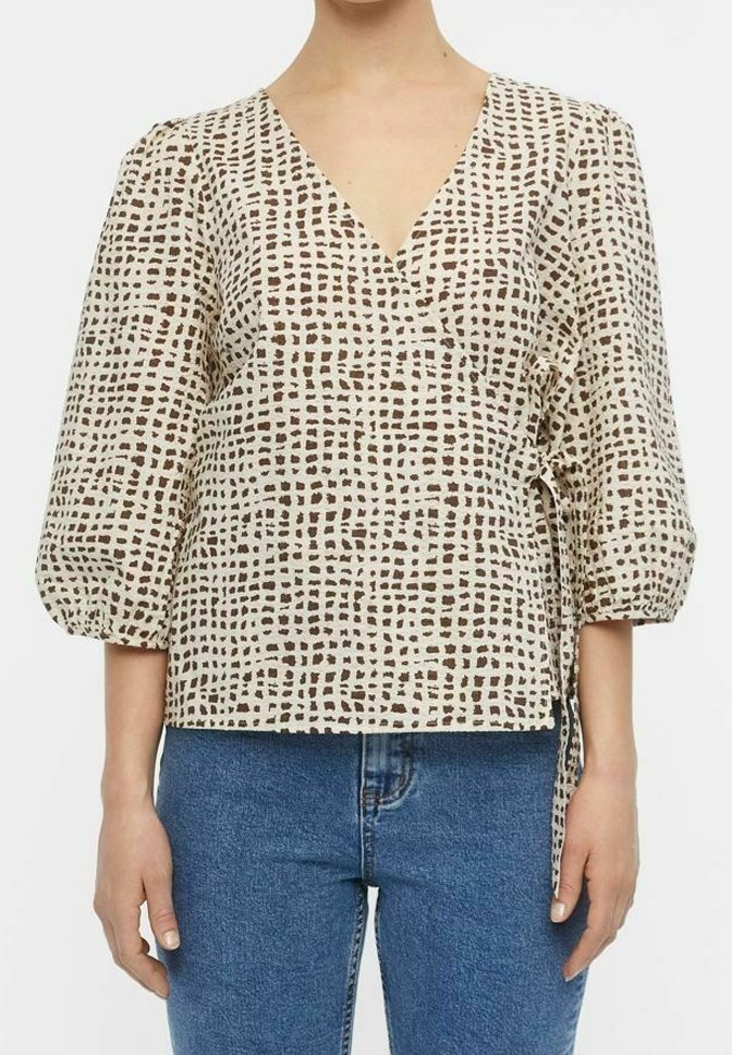 Top beige enroulé avec un décolleté en V, motif en grille texturé marron, manches bouffantes et un lien sur le côté, associé à un jean en denim bleu.