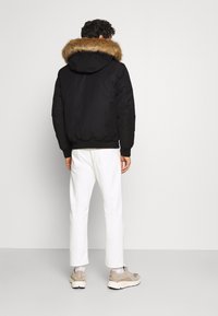 Schwarze Kapuzenjacke mit einem Kragen aus Kunstfell, elastischem Bund und Bündchen, kombiniert mit weißen Hosen und beigen Sneakers, von hinten betrachtet.