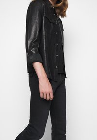 Camisa de cuero negro con un diseño ligeramente oversize, que incluye cierres de botones y dos bolsillos en el pecho, combinada con jeans negros de corte slim.