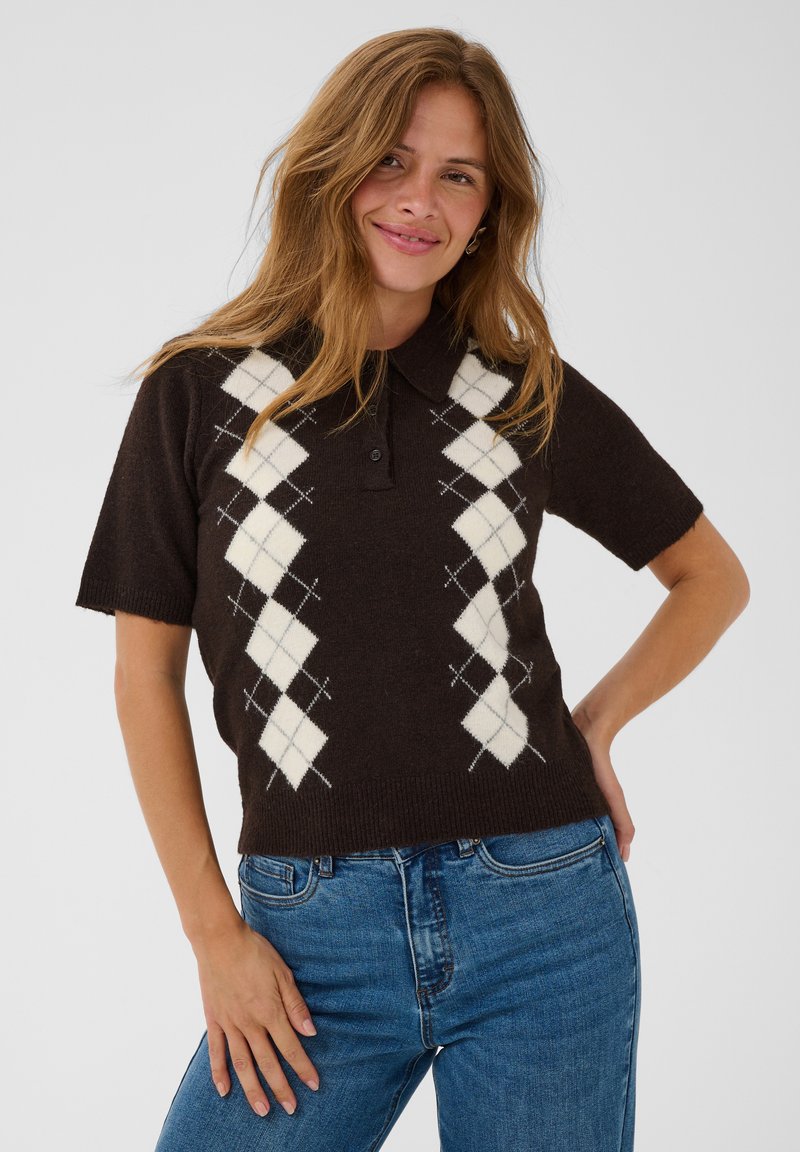 Femme aux longs cheveux châtain clair porte un pull foncé à losanges à manches courtes et un jean bleu, souriante, une main sur la hanche devant un fond uni.
