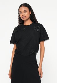 Top nero a maniche corte in tessuto testurizzato, con decorazioni floreali in paillettes sul davanti. Vestibilità morbida e rilassata con scollatura rotonda.