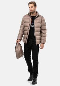 Puffers akte in hellbraun mit hohem Kragen und horizontalen Nähten. Modell hält einen passenden leichten Rucksack. Schwarze Hose und Schuhe.