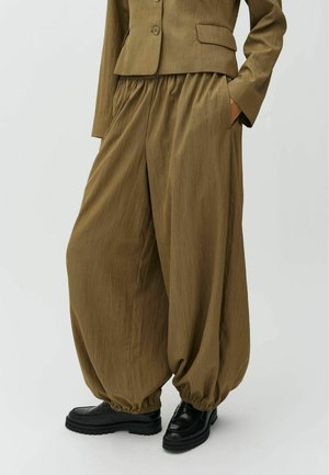 Pantaloni marroni larghi con polsini elastici abbinati a una giacca marrone coordinata e scarpe nere robuste su sfondo bianco.