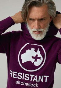 Sudadera con capucha morada con un gráfico blanco que muestra un contorno de mapa y texto que dice "RESISTANT" con el nombre de la marca "altonadock" debajo. Textura suave.