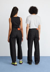 Svart bredbente jeans med oransje baketiketter blir brukt av to personer, en i en svart tanktop og den andre i en grå t-skjorte.