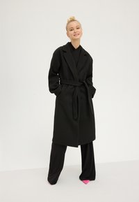 Blonde woman wearing a long black belted coat, black wide-leg pants, and pink pointed shoes, standing against a plain light background.