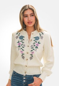 Mujer con un suéter crema abotonado con bordado floral en azul y rosa, combinado con jeans de cintura alta azul y joyas doradas.