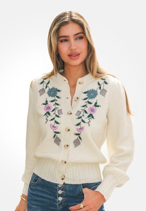 Mujer con un suéter crema abotonado con bordado floral en azul y rosa, combinado con jeans de cintura alta azul y joyas doradas.