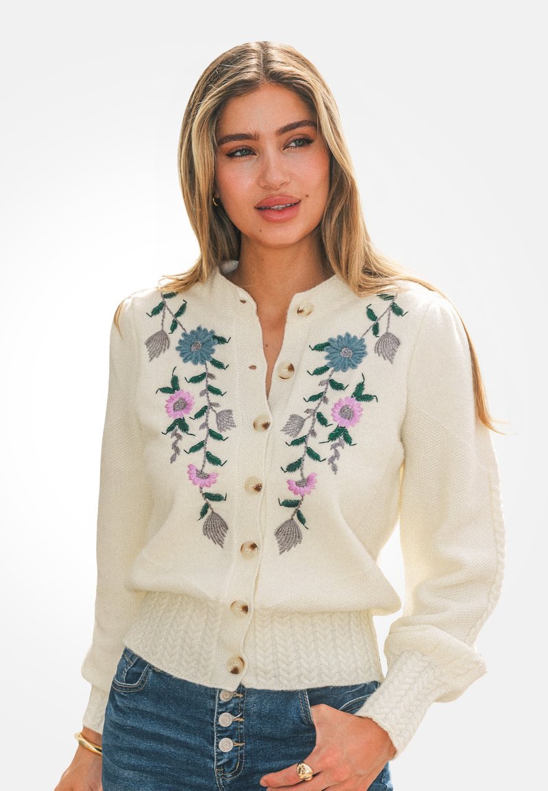 Mujer con un suéter crema abotonado con bordado floral en azul y rosa, combinado con jeans de cintura alta azul y joyas doradas.