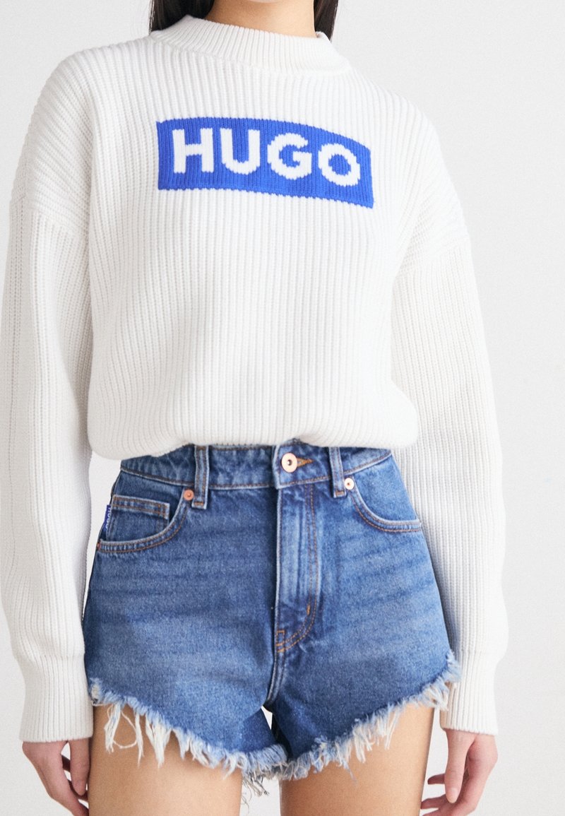 Maglione bianco a coste con un logo "HUGO" blu e bianco, abbinato a pantaloni shorts in denim blu sfilacciati con hardware rame e vita alta.