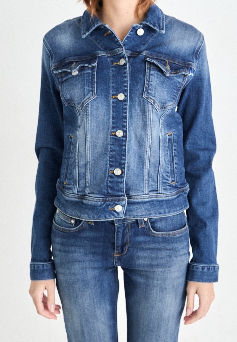 Femme portant une veste en denim bleue avec fermeture par boutons et un jean denim bleu assorti, debout devant un fond clair uni.