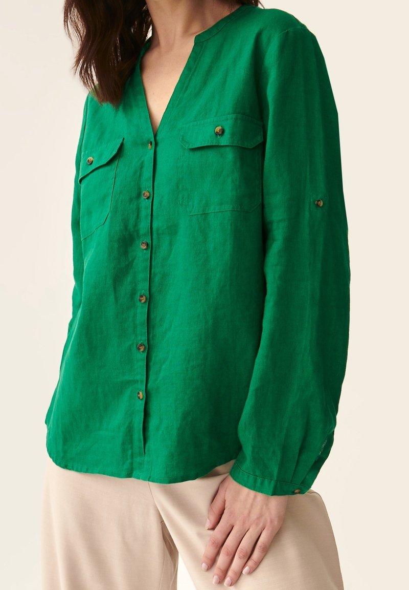 Femme portant une chemise boutonnée à manches longues vert vif avec deux poches poitrine et un pantalon beige, la main reposant sur la cuisse.