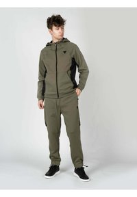 Chándal con capucha de color verde oliva con paneles laterales negros y bolsillos con cremallera; presenta puños acanalados y cintura ajustable; combinado con zapatillas negras.