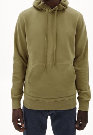 Personne portant un sweat à capuche vert olive avec poche frontale et cordons, associé à un jean bleu foncé sur un fond uni.