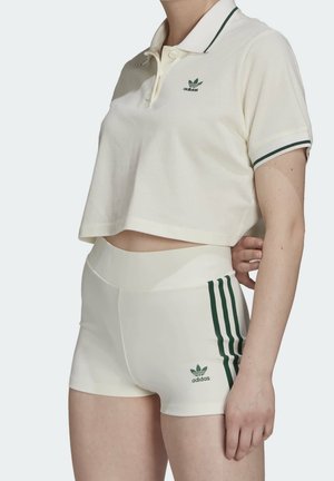 Femme portant un polo Adidas écru court et un short taille haute assorti avec trois bandes vertes et logo sur le côté gauche.