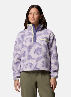 Frau trägt einen lavendelfarbenen und violetten floralen Fleece-Pullover mit Druckknöpfen und einer Reißverschlusstasche auf der Brust, kombiniert mit olivgrüner Hose.