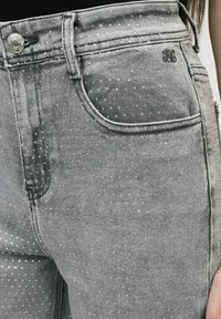 Gros plan sur un jean en denim gris avec de petits pois blancs, poche avant, bouton en métal et passants de ceinture, porté par une personne.