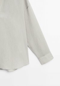 Manche de chemise à manches longues de couleur beige clair avec fines rayures verticales, montrant le poignet et une partie du corps de la chemise sur un fond blanc.
