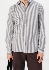 Raidallinen paita vaaleanharmaana ja tummansinisena, jossa on napillinen kaulus, pitkät hihat ja suora helma kapeilla hihansuiden päällisillä.