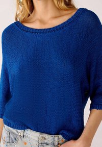 Niebieski sweter dzianinowy z szerokim dekoltem i rękawami o długości trzy czwarte, z teksturowanym wzorem i luźnym krojem. Zestawiony z jasnymi dżinsowymi szortami.