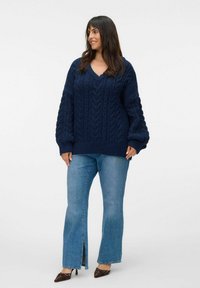 Femme souriante, portant un pull en tricot torsadé bleu marine, un jean bleu clair avec fentes à l'avant et des escarpins pointus noirs devant un fond uni.